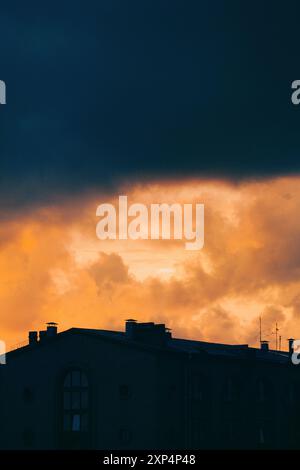 La silhouette sombre du toit du bâtiment de la ville est faiblement éclairée par la lueur orange du soleil couchant, au-dessus duquel pendent de lourds nuages de tonnerre. Banque D'Images