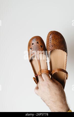 Main masculine présentant une paire de sandales d'été de luxe pour femmes en cuir perforé sur un fond blanc, soulignant le design élégant et l'artisanat de haute qualité Banque D'Images