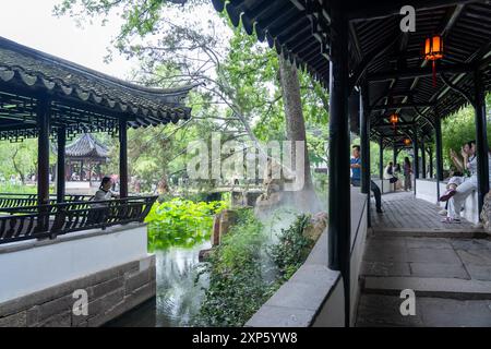 Suzhou, Chine - 11 juin 2024 : Un jardin chinois traditionnel à Suzhou, avec une passerelle couverte, un étang avec des nénuphars et un pavillon dans le dist Banque D'Images