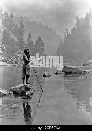 Une journée enfumée au Sugar Bowl--Hupa. Homme Hupa avec lance, debout sur la roche à mi-courant, en arrière-plan, le brouillard obscurcit partiellement les arbres sur les flancs des montagnes. Publié en 1924 dans The North American Indian / E Banque D'Images