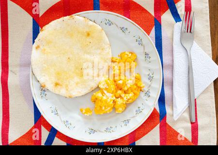 Une photo aérienne présentant un petit-déjeuner colombien typique, avec une assiette avec arepa et œufs brouillés sur une nappe colorée et vibrante Banque D'Images