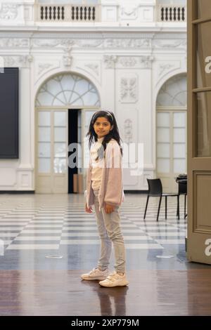 Une jeune fille se tient dans une grande pièce avec un fond blanc. Elle porte un pull rose et un pantalon gris Banque D'Images