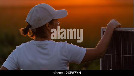 Femme portant un chapeau blanc se tient à côté du panneau solaire au coucher du soleil. Banque D'Images