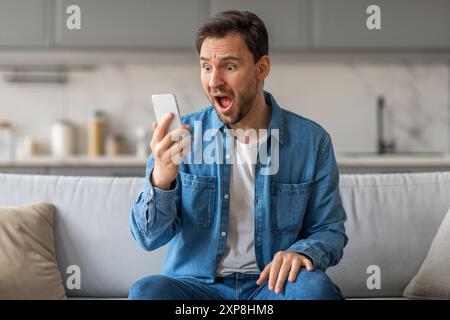 Un homme choqué assis sur un canapé regarde son smartphone Banque D'Images