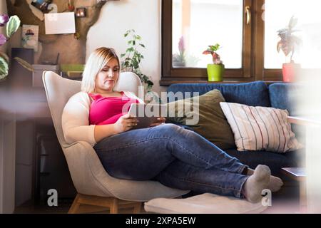 Portrait de belle jeune femme en surpoids à la maison, regardant l'émission de télévision sur tablette, assis dans un fauteuil. Banque D'Images