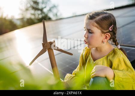 Fille mignonne sur le toit avec des panneaux solaires, soufflant dans le modèle de l'éolienne. Installation solaire ou photovoltaïque sur le toit. Un avenir durable pour la prochaine génération. Banque D'Images