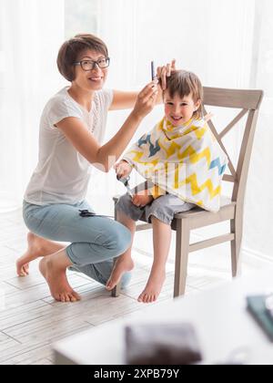 Mère coupe les cheveux de son fils par elle-même. Le petit garçon est assis, recouvert de tissu, et tient une paire de ciseaux. Nouveau normal en cas de coronavirus COVID-19 qua Banque D'Images