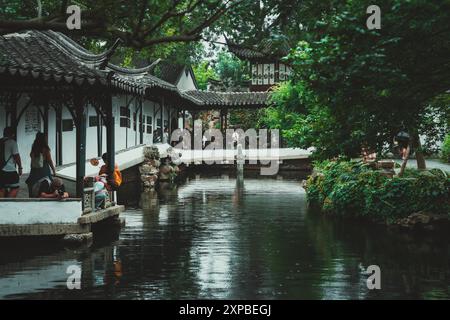 Suzhou, Chine - 11 juin 2024 : Un jardin chinois tranquille avec un étang, un pont de pierre et un bâtiment traditionnel avec un toit incurvé. Banque D'Images