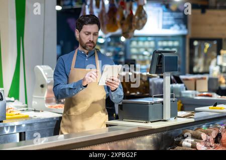 Ouvrier d'épicerie masculin utilisant la tablette à la caisse. Travailleur portant tablier, debout dans un supermarché moderne, vérifiant l'inventaire et les prix sur l'appareil. Banque D'Images