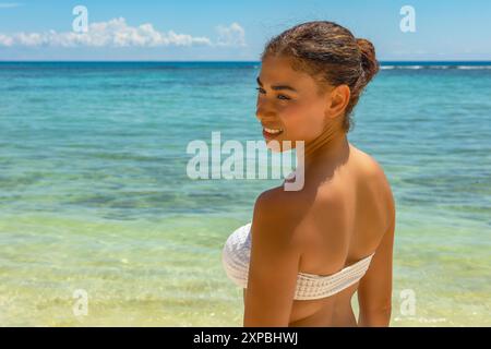 Une tempête tropicale trouve son bonheur sur une plage isolée. Vêtue d'un bikini blanc, elle respire la confiance et l'allure sur fond de turquoise w Banque D'Images