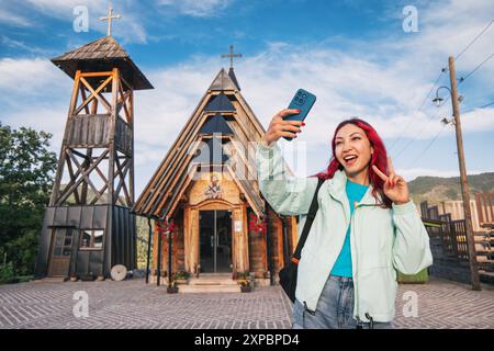 10 juin 2024, Mokra Gora, Serbie : femme voyageant à Drvengrad, capturant un selfie avec son smartphone devant une belle maison en bois Banque D'Images