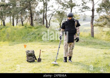 Un homme dans un uniforme militaire et gilet à l'épreuve des balles travaille dans la forêt avec un détecteur de métaux. Un mineur effectue des travaux de déminage du territoire. Banque D'Images