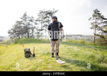 Un homme dans un uniforme militaire et gilet à l'épreuve des balles travaille dans la forêt avec un détecteur de métaux. Un mineur effectue des travaux de déminage du territoire. Banque D'Images