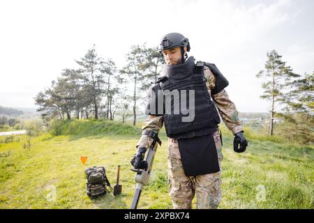 Un homme dans un uniforme militaire et gilet à l'épreuve des balles travaille dans la forêt avec un détecteur de métaux. Un mineur effectue des travaux de déminage du territoire. Banque D'Images