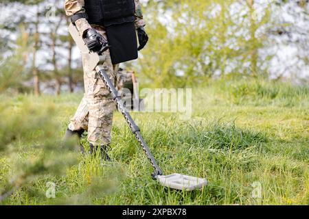 Un homme dans un uniforme militaire et gilet à l'épreuve des balles travaille dans la forêt avec un détecteur de métaux. Un mineur effectue des travaux de déminage du territoire. Banque D'Images
