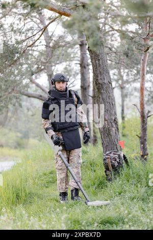 Un homme dans un uniforme militaire et gilet à l'épreuve des balles travaille dans la forêt avec un détecteur de métaux. Un mineur effectue des travaux de déminage du territoire. Banque D'Images