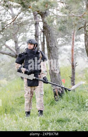 Un homme dans un uniforme militaire et gilet à l'épreuve des balles travaille dans la forêt avec un détecteur de métaux. Un mineur effectue des travaux de déminage du territoire. Banque D'Images