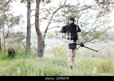 Un homme dans un uniforme militaire et gilet à l'épreuve des balles travaille dans la forêt avec un détecteur de métaux. Un mineur effectue des travaux de déminage du territoire. Banque D'Images