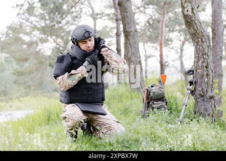 Un homme dans un uniforme militaire et un gilet à l'épreuve des balles travaille dans la forêt pour déminer le territoire. Un homme met un casque de protection avant de commencer le travail Banque D'Images