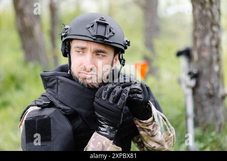 Un homme dans un uniforme militaire et un gilet à l'épreuve des balles travaille dans la forêt pour déminer le territoire. Un homme met un casque de protection avant de commencer le travail Banque D'Images