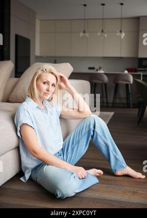 Magnifique femme assise à côté du canapé dans son appartement. Jolie femme dans des vêtements décontractés regardant droit sur la caméra. Banque D'Images