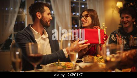 Beau petit ami présentant un cadeau festif à sa belle petite amie surprise. Famille et amis se réunissent à la maison dans la soirée pour un dîner de Noël traditionnel avec un rôti de dinde Banque D'Images