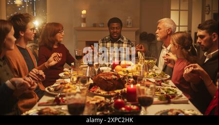 Portrait d'un jeune homme noir assis à une table de fête avec famille et amis, priant avant de manger un dîner de dinde. Religieux Africain bénissant Noël nourriture, remerciant Dieu et Jésus Christ Banque D'Images