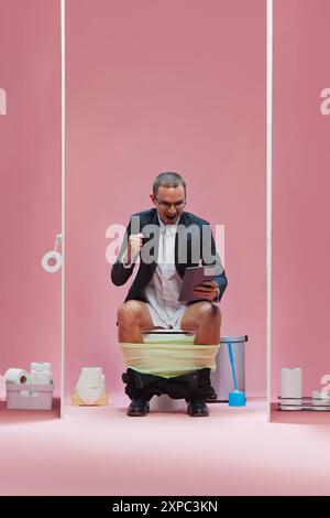 Homme d'affaires en costume assis sur les toilettes avec pantalon vers le bas et regardant émotionnellement la traduction de jeu de football en ligne via tablette sur fond rose Banque D'Images