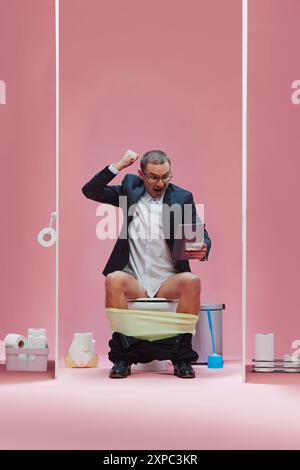 Objectif. Homme émotionnel en vêtements formels assis avec un pantalon vers le bas sur les toilettes et regardant le match de football via la tablette et criant sur fond rose Banque D'Images