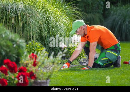 Un jardinier dans une chemise orange et un chapeau vert s'agenouille à côté de fleurs vibrantes, s'occupant soigneusement des plantes dans un jardin bien entretenu. Banque D'Images