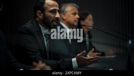 Chef de délégation politique masculin multiethnique prenant la parole lors de la conférence de presse du Forum économique international. Politicien de haut niveau qui rend compte des accords réussis. Différents participants écoutent. Banque D'Images