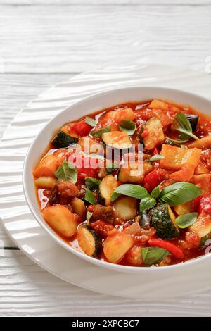 Ciambotta, Giambotta, ragoût de légumes italiens de courgettes, aubergines, poivrons, pomme de terre, basilic frais et sauce tomate dans un bol blanc sur t en bois blanc Banque D'Images