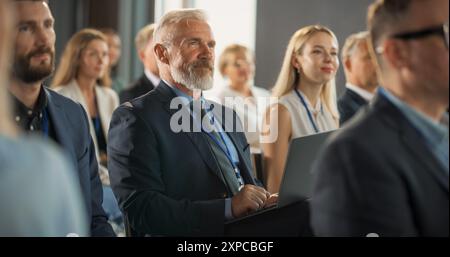 Investisseur masculin senior assis dans la foule et utilisant un ordinateur portable à la Conférence internationale sur la technologie. Homme caucasien écoutant la présentation Keynote sur Innovative Startup Company Service. Banque D'Images