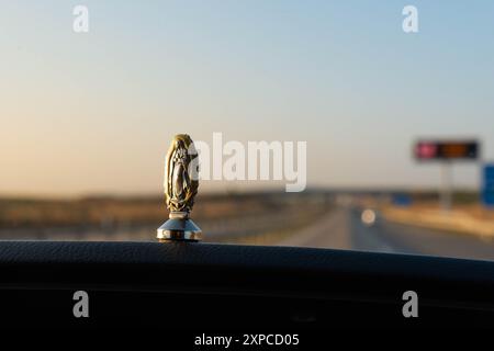 Une figurine de la Vierge de Guadalupe collée sur le tableau de bord d'une voiture. En arrière-plan, une autoroute hors foyer avec un grand pont un panneau. Christia Banque D'Images