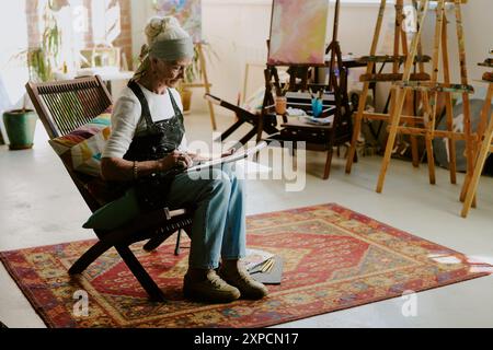 Artiste caucasien senior portant tablier assis sur une chaise en bois dans son studio d'art et dessinant dans un carnet de croquis, espace copie Banque D'Images