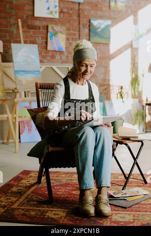 Long plan vertical de femme caucasienne âgée portant un tablier assis sur une chaise dans un studio d'art et dessinant dans un carnet de croquis Banque D'Images