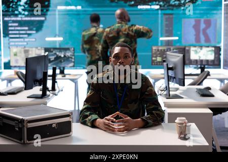 Membre de l'équipage militaire participant à une connexion vidéo en ligne sécurisée, intégrant des données en temps réel et une technologie de surveillance avancée dans un centre de commandement pour des opérations stratégiques. Banque D'Images