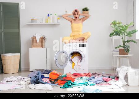 Jeune femme choquée assise sur la machine à laver avec des piles de vêtements sales sur le sol dans la buanderie Banque D'Images