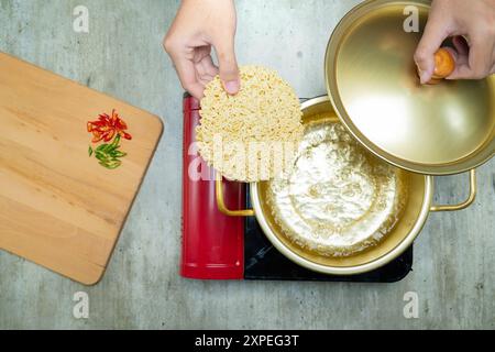 Une personne fait cuire des nouilles instantanées dans une casserole avec un couvercle sur le dessus. Le pot est sur un poêle et il y a une planche à découper en bois à proximité Banque D'Images