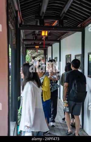 Suzhou, Chine - 11 juin 2024 : Un groupe de personnes se promène dans un jardin chinois traditionnel à travers une passerelle couverte avec un plafond en bois et des murs blancs. Banque D'Images