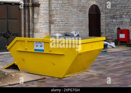 Une benne à ordures jaune vif se dresse près d'un bâtiment en pierre, entourée de débris et de matériaux de construction dans un environnement urbain animé. Banque D'Images