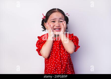 Jeune fille asiatique souriant joyeusement avec les mains sur les joues, portant une robe à pois rouges et des rubans rouges et blancs sur les cheveux Banque D'Images