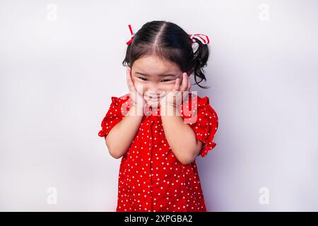 Jeune fille asiatique, souriant avec les deux mains sur les joues, portant une robe à pois rouges et des rubans rouges et blancs sur les cheveux isolés sur fond blanc. Banque D'Images