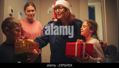 Jeune mère rencontre Grand-parent par la porte dans une maison décorée avec des guirlandes, petit garçon et fille courir pour rencontrer leur grand-mère avec des cadeaux de Noël et des cadeaux Banque D'Images