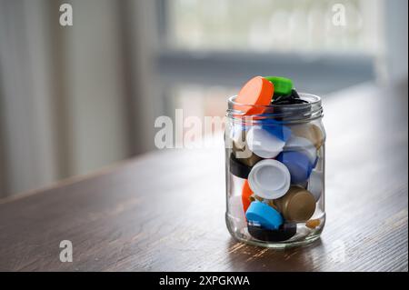 Pot en verre se trouve sur une table en bois, remplie de bouchons de bouteille en plastique coloré. Les bouchons sont collectés pour être recyclés Banque D'Images