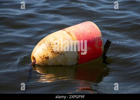 Gros plan d'une bouée altérée marquée de bandes rouges et jaunes flotte doucement sur une eau calme, attachée pour indiquer une zone de navigation ou de sécurité. Banque D'Images