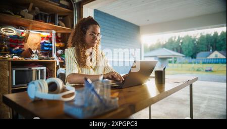 Heureuse femme hispanique utilisant l'ordinateur portable dans le garage ouvert. Entrepreneur numérique féminin réussi exécutant une startup de commerce électronique à la maison, vérifiant les e-mails, remplissant les commandes en ligne, recherchant le marché. Banque D'Images
