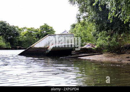 Un vieux bateau partiellement submergé repose sur une rive, appuyé contre la rive boueuse. Une végétation dense entoure la région, avec des arbres bordant la rivière Banque D'Images