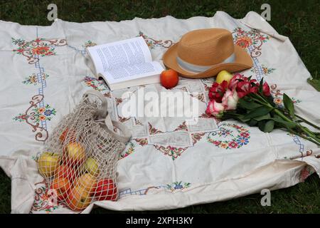 Bouquet de roses, pommes, livre ouvert et chapeau de paille sur une couverture de pique-nique. Activités du week-end d'été. Concept de vie saine. Banque D'Images