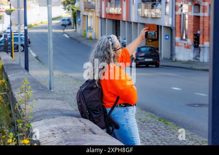 Touriste féminine prenant un selfie, appuyée sur une clôture sur un trottoir à côté d'une rue urbaine, bâtiments en arrière-plan flou, journée ensoleillée lors d'une visite à DIN Banque D'Images